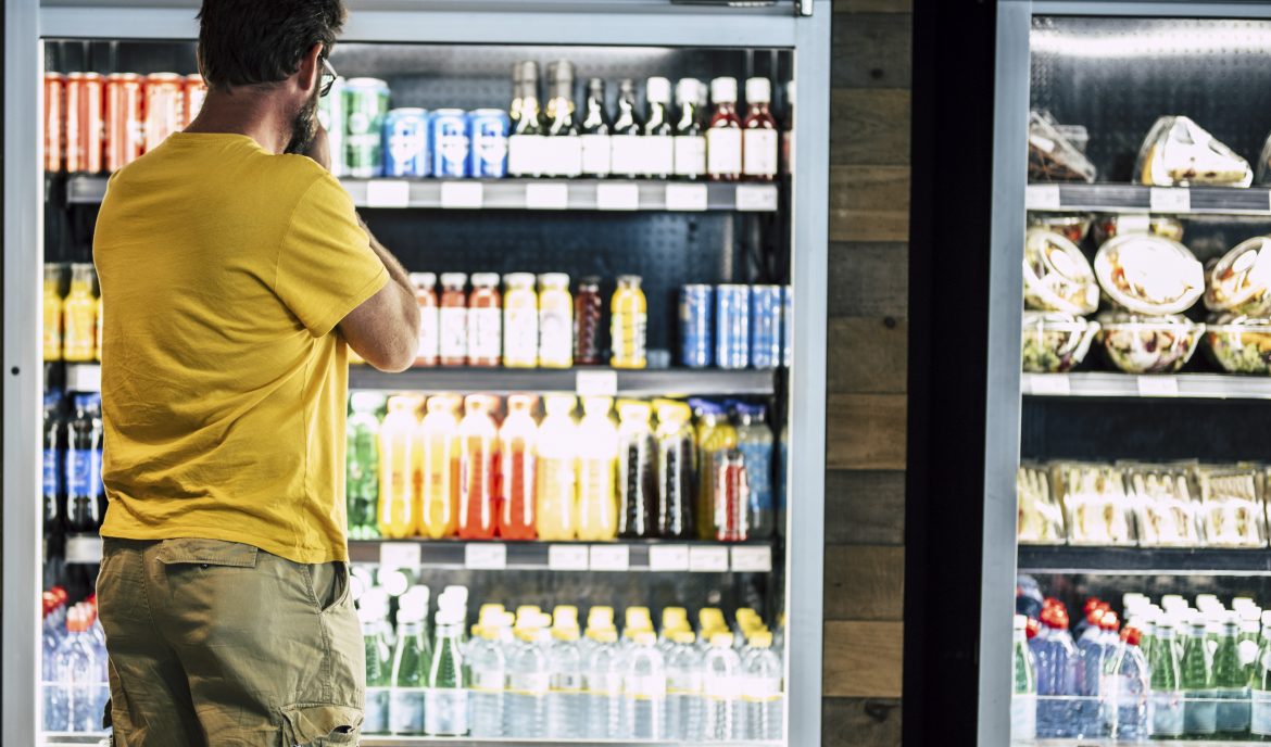 man alone and isolated at the supermarket or minimarket choosing his drink or snack in big fridge with lots of products - difficult select and choose